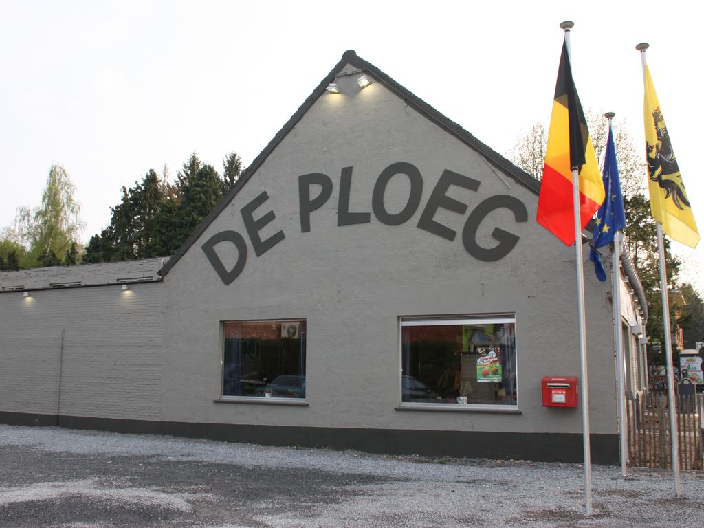 De gevel van Vrijetijdscentrum De Ploeg aan de Kapellei: een lage witte zaal met “DE PLOEG” in grote letters op de puntgevel, met de Belgische, Europese en Vlaamse vlag ervoor en parking op de voorgrond.
